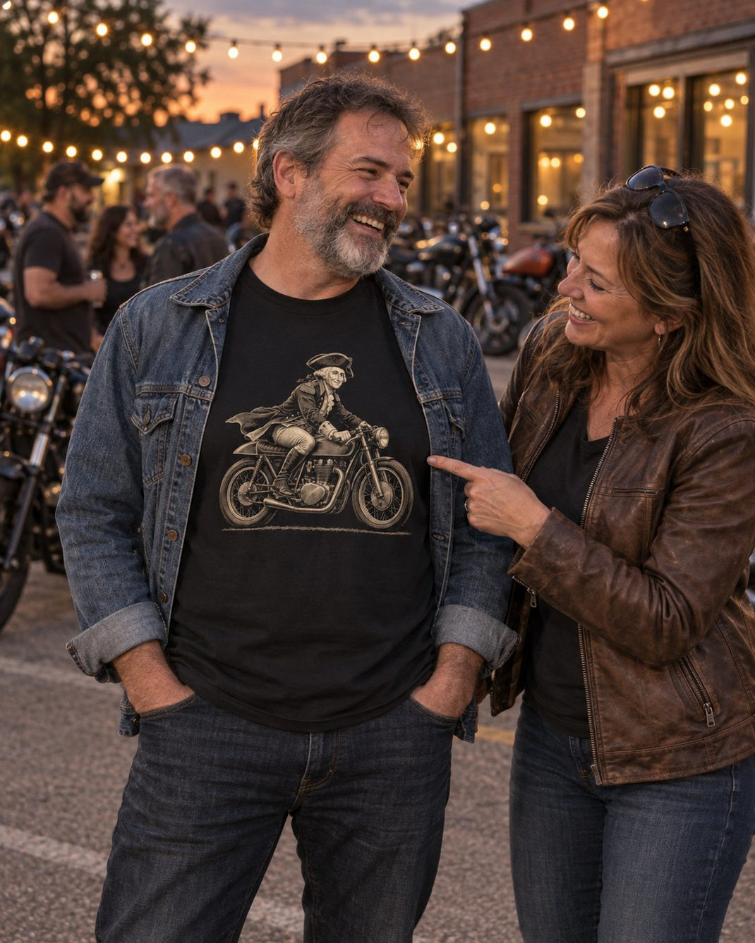 At a dusk bike night, a woman laughs and points at the colonial rider on a cafe racer printed on a man's black tee
